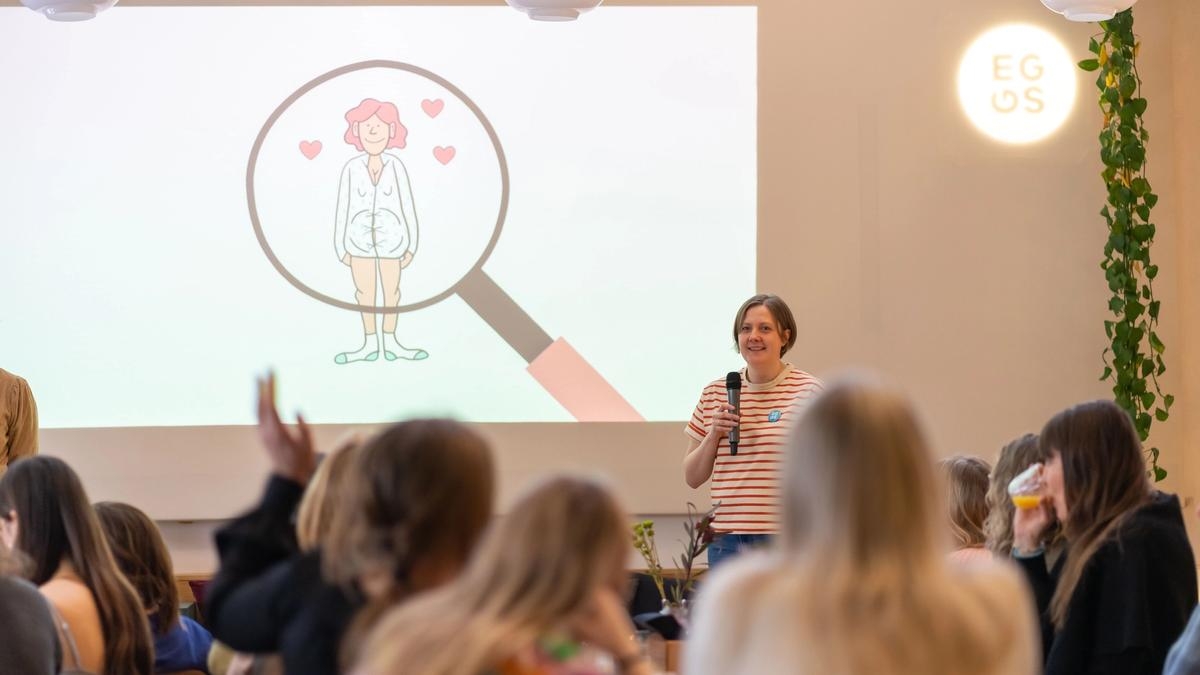 Designer Gøril Storrø stands in front of an audience with a microphone in her hand and behind her is a power point projection of an illustrated, pregnant woman. Photo.