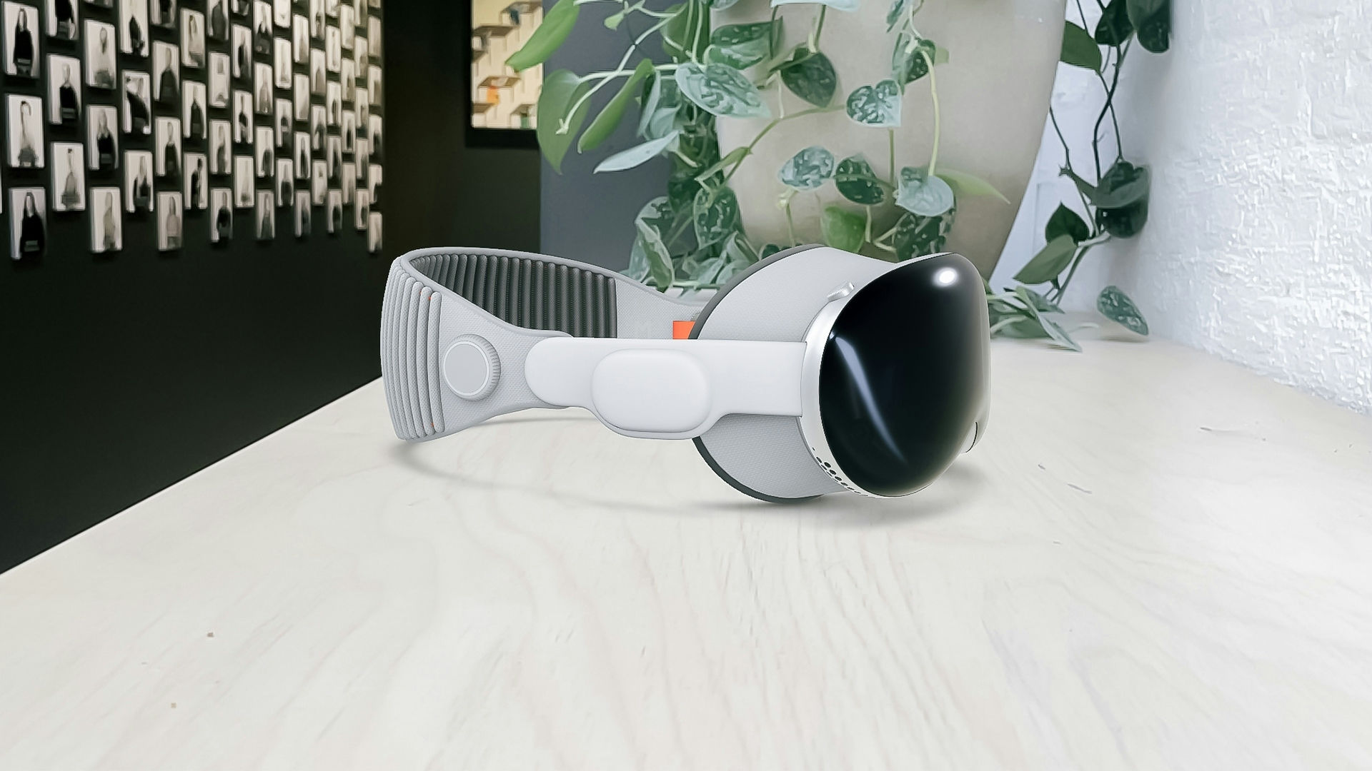 An Apple Vision Pro device placed on a wooden surface. A plant and dark wall with photos showing in the background. Photo and render.